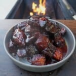 A plate of finished honey chipotle pork belly burnt ends, swathed in sauce and sitting on a table in front of a grill fire