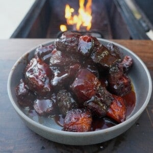 A plate of finished honey chipotle pork belly burnt ends, swathed in sauce and sitting on a table in front of a grill fire