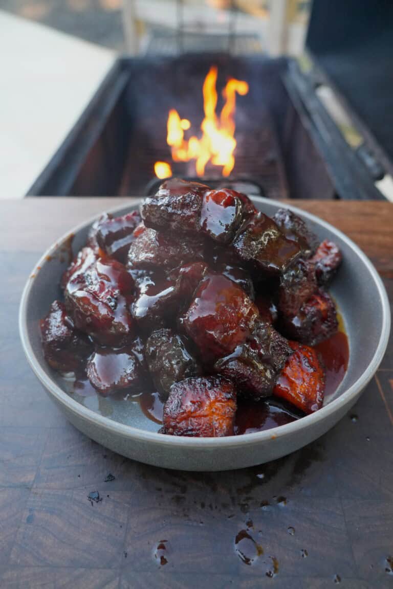 A plate of finished honey chipotle pork belly burnt ends, swathed in sauce and sitting on a table in front of a grill fire