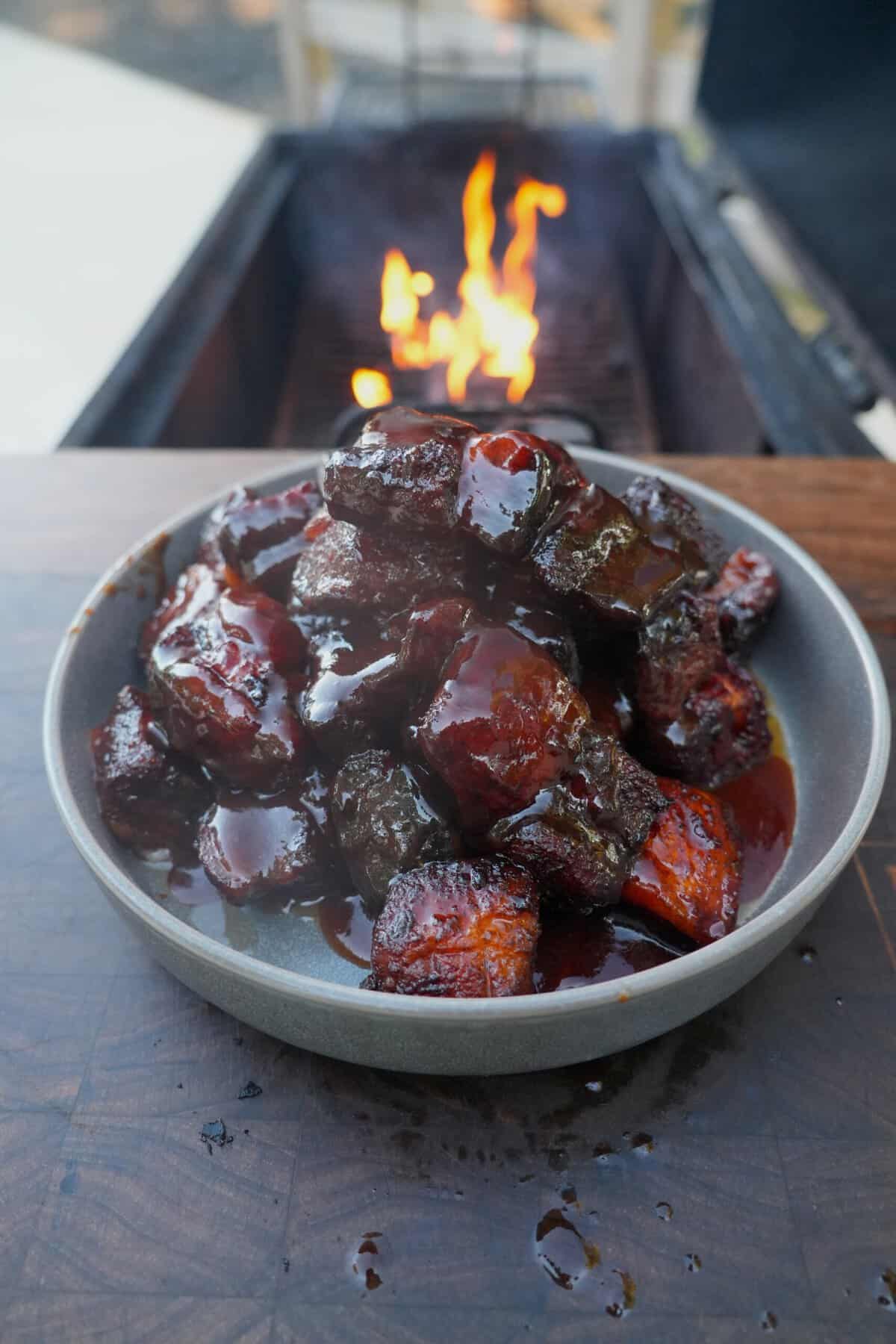 A plate of finished honey chipotle pork belly burnt ends, swathed in sauce and sitting on a table in front of a grill fire